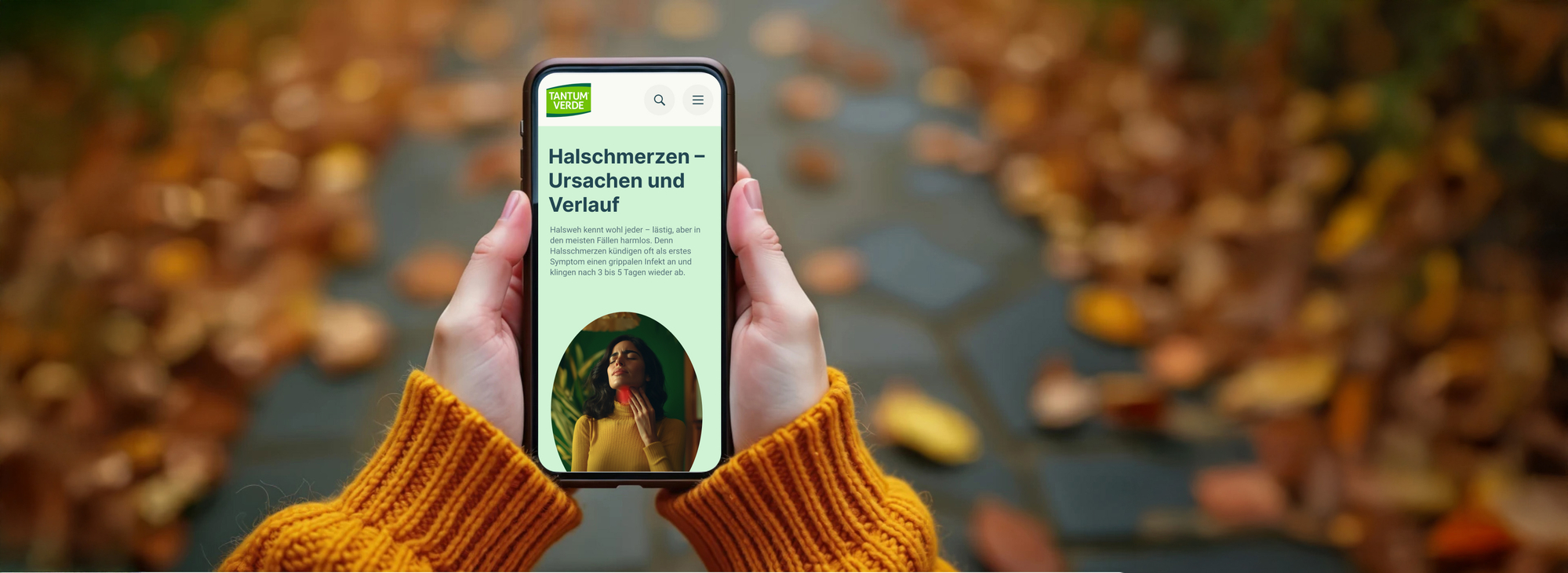 Person hält ein Smartphone mit einer Gesundheitswebseite zum Thema „Halsschmerzen – Ursachen und Verlauf“ in den Händen; im Hintergrund ein herbstlicher Weg mit buntem Laub.