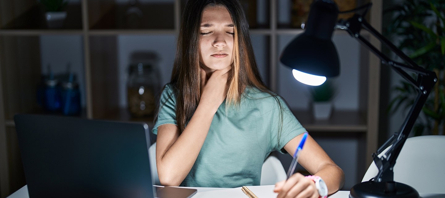 Ein Mädchen hält sich den Hals vor Schmerzen, während sie vor ihrem Laptop am Schreibtisch sitzt und lernt