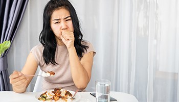 Ansicht einer jungen Frau mit schmerzverzerrtem Gesicht am Esstisch, die eine volle Gabel hält und sich gleichzeitig die andere, zur Faust geballte Hand vor den Mund hält.