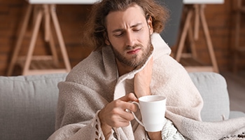 Ein Mann sitzt mit einer Decke auf der Couch, fasst sich den Hals vor Schmerzen und hält eine Teetasse in der Hand