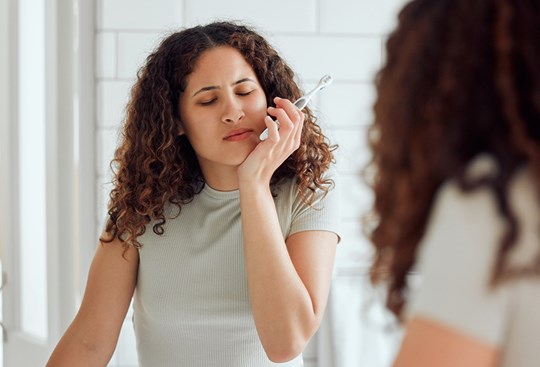 Eine junge Frau hält sich beim Zähneputzen vor dem Badezimmerspiegel mit der Hand die schmerzende Wange. 