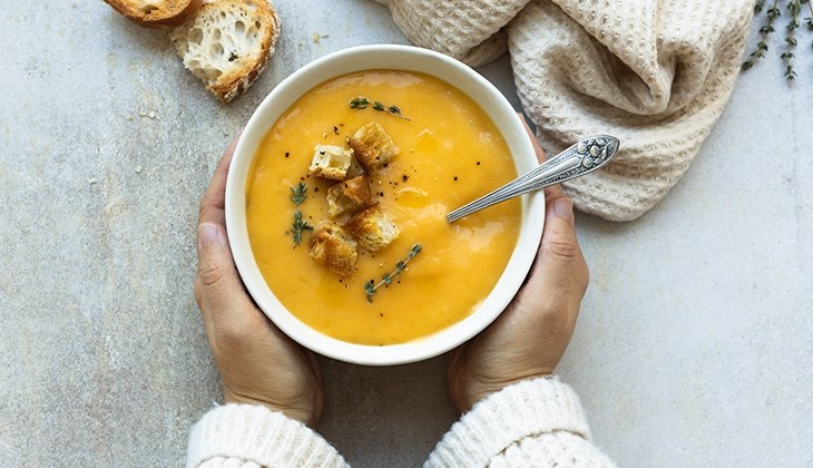 Person hält eine Schüssel Kürbissuppe mit Croutons und frischen Kräutern, daneben liegen Brotscheiben.