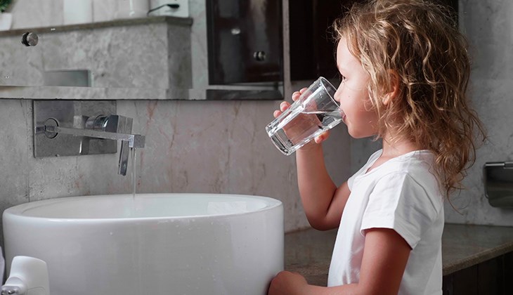 Seitliche Ansicht eines kleinen Kindes, das vor einem Badezimmerwaschbecken steht und ein Glas Wasser trinkt.