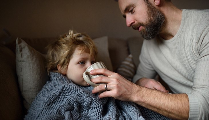 Ein Vater gibt seinem kranken Sohn, der in eine Deckel eingewickelt ist, Tee
