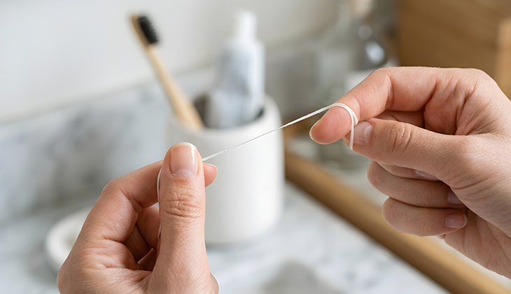 Zwei Hände halten Zahnseide zwischen Daumen und Zeigefinger. Im Hintergrund Becher mit Zahnbürste und Zahnpasta.