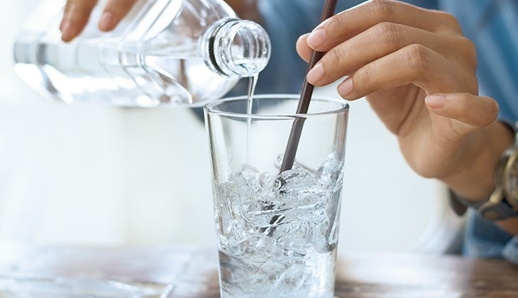 Eine Person gießt sich Wasser aus einer Flasche in ein Glas mit Eiswürfeln und Strohhalm ein