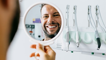 Ein Mann hält in einer Zahnarztpraxis einen Handspiegel hoch. Der Spiegel zeigt sein lächelndes Gesicht mit weißen Zähnen.