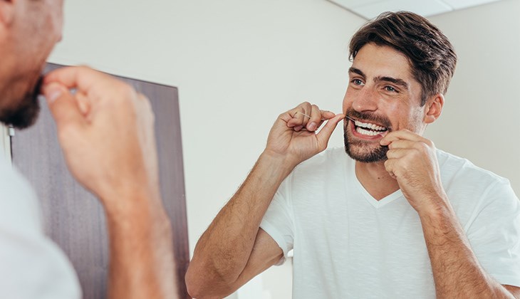 Eine lächelnder junger Mann verwendet Zahnseide vor dem Badezimmerspiegel.
