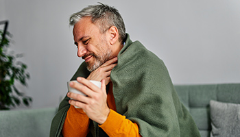 Ein Mann mit einer Teetasse in der Hand sitzt in eine Decke eingehüllt auf dem Sofa und greift sich mit schmerzverzerrtem Gesicht an den Hals