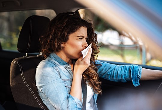 Eine dunkelhaarige Frau sitzt im Auto und fährt, dabei hustet sie und hält sich ein Taschentuch vor den Mund