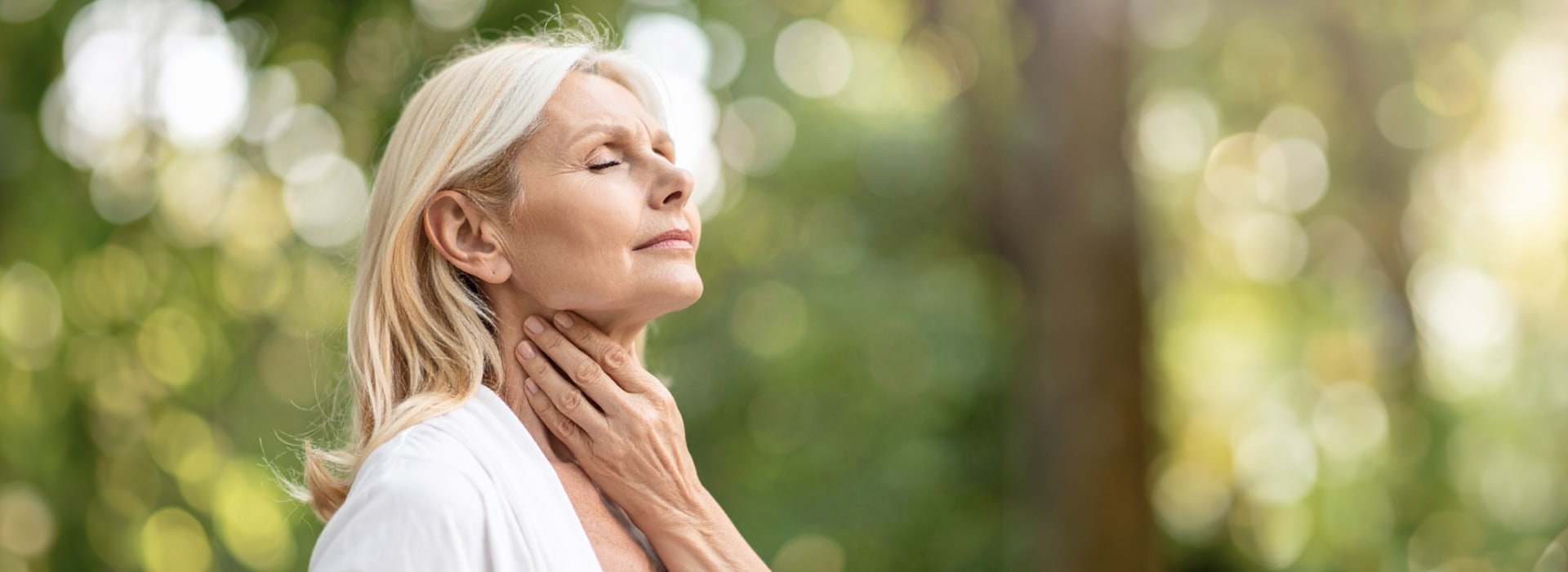 Ältere Frau mit Halsschmerzen fasst sich an den Hals und steht entspannt im Grünen.