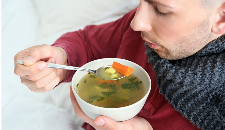 Mann mit Schal hält eine Schale Gemüsesuppe und löffelt daraus.
