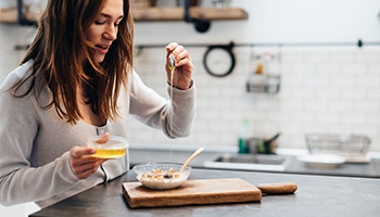 Eine junge Frau steht in einer modernen Küche und gießt mit einem Löffel Honig über eine Schüssel Müsli.