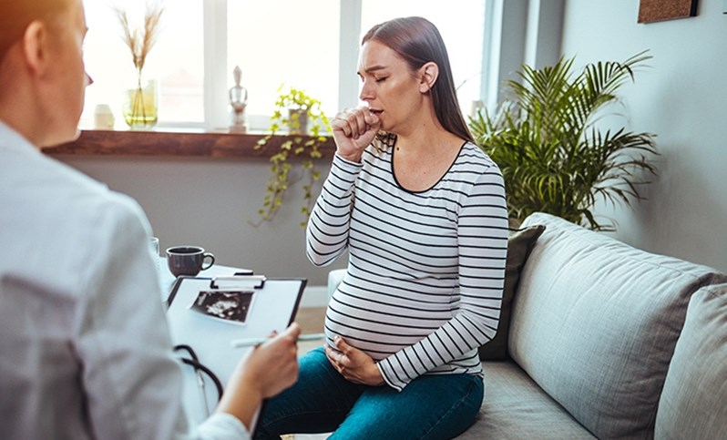 Eine schwangere Frau sitzt in einer Arztpraxis und hustet