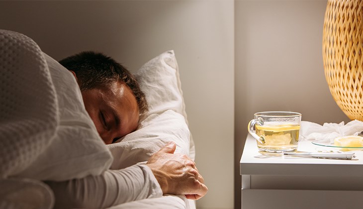 Kranker Mann schläft im abgedunkelten Schlafzimmer; auf dem Nachttisch stehen ein Tee mit Zitrone, Tabletten und Taschentücher.