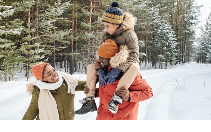 Mutter, Vater und Kind laufen einen verschneiten Waldweg lang, während das Kind auf den Schultern vom Vater sitzt