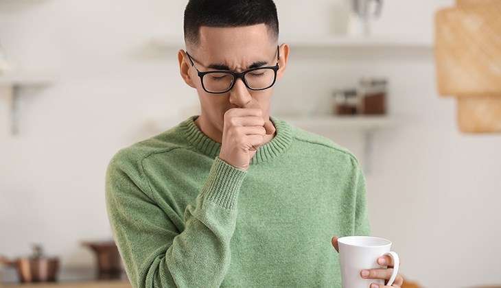 Ein Mann in gruenem Pullover hustet in seine rechte Hand und haelt mit der linken Hand eine Tasse Tee