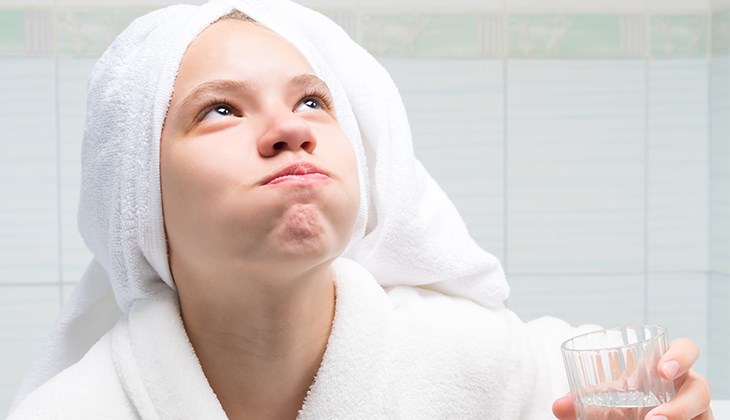 Junge Person im Bademantel mit Handtuch-Turban gurgelt mit einem Glas Wasser bzw. einer Mundspülung.