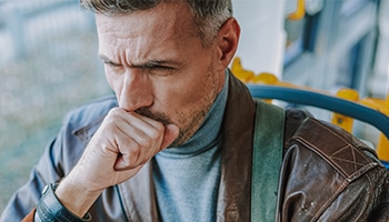 Ein Mann mit grauen Haaren sitzt im Bus, hustet und hält sich dabei die Hand vor den Mund
