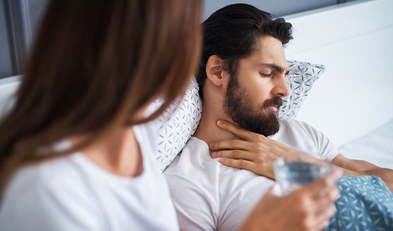 Ein Mann liegt mit Halsschmerzen im Bett, eine Frau umsorgt ihn mit Wasser