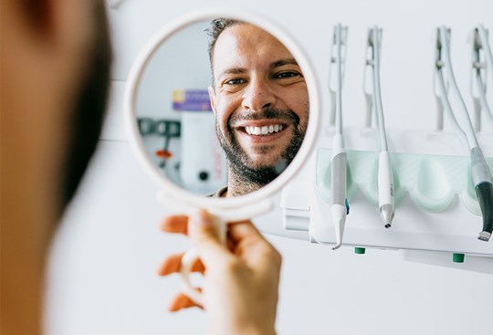 Ein Mann hält in einer Zahnarztpraxis einen Handspiegel hoch. Der Spiegel zeigt sein lächelndes Gesicht mit weißen Zähnen.