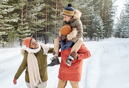 Mutter, Vater und Kind laufen einen verschneiten Waldweg lang, während das Kind auf den Schultern vom Vater sitzt
