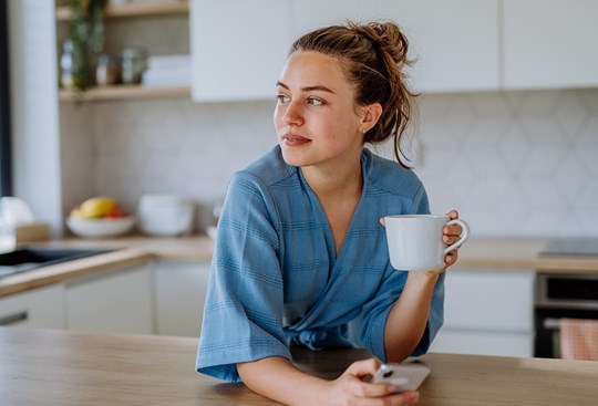 Frau mit Dutt steht im Morgenmantel auf die Küchentheke gelehnt und hält eine Tasse Tee in der linken Hand