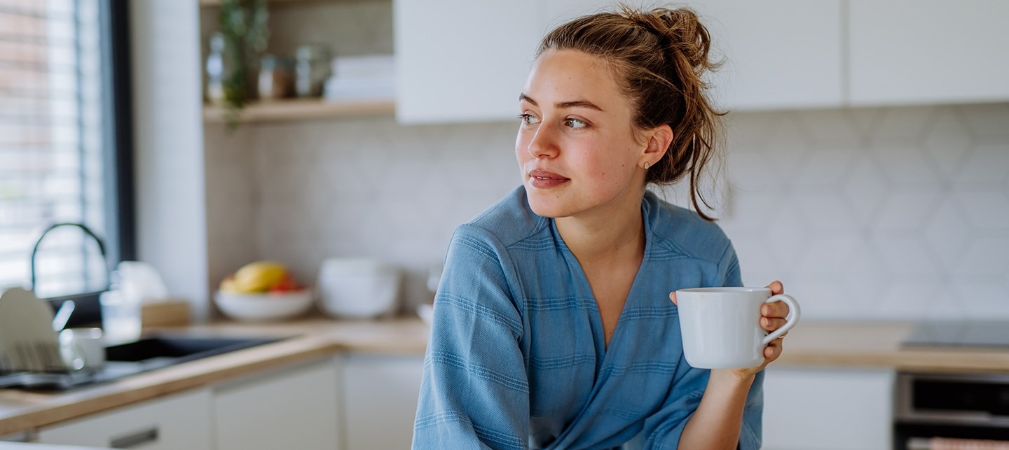 Frau mit Dutt steht im Morgenmantel auf die Küchentheke gelehnt und hält eine Tasse Tee in der linken Hand