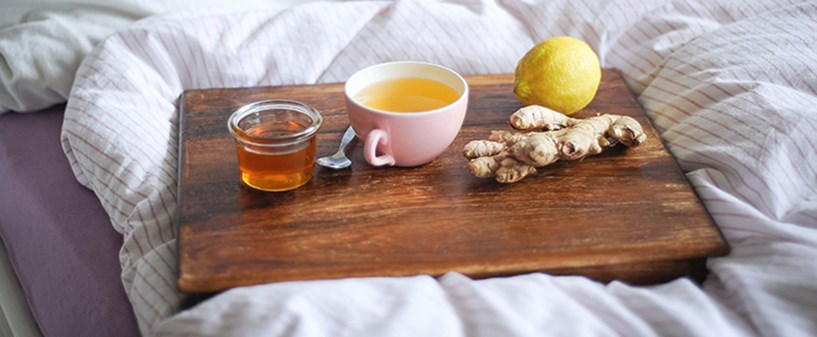 Auf einem Tablett steht eine Tasse Tee und ein Glas Honig umgeben von Ingwer und Zitrone