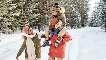 Mutter, Vater und Kind laufen einen verschneiten Waldweg lang, während das Kind auf den Schultern vom Vater sitzt