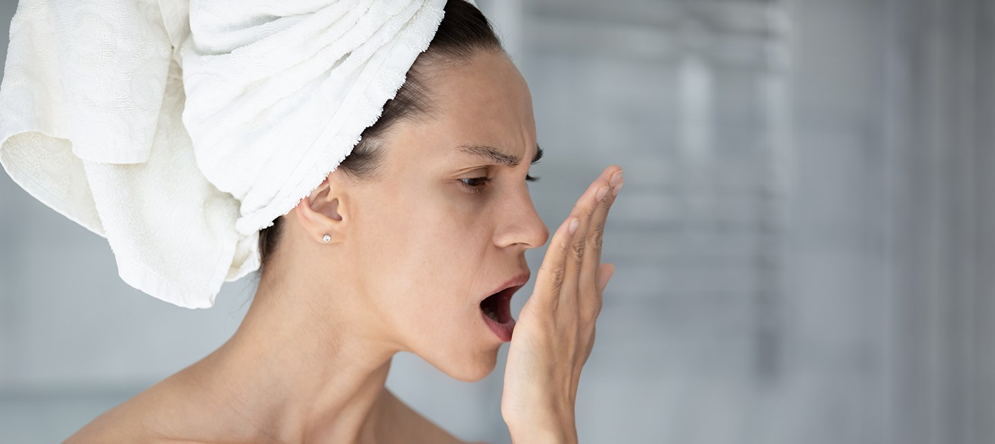 Halbseitliche Kopf- und Schulteransicht einer Frau mit Handtuchturban, die mit kritischem Blick ihre eigene Hand anhaucht.