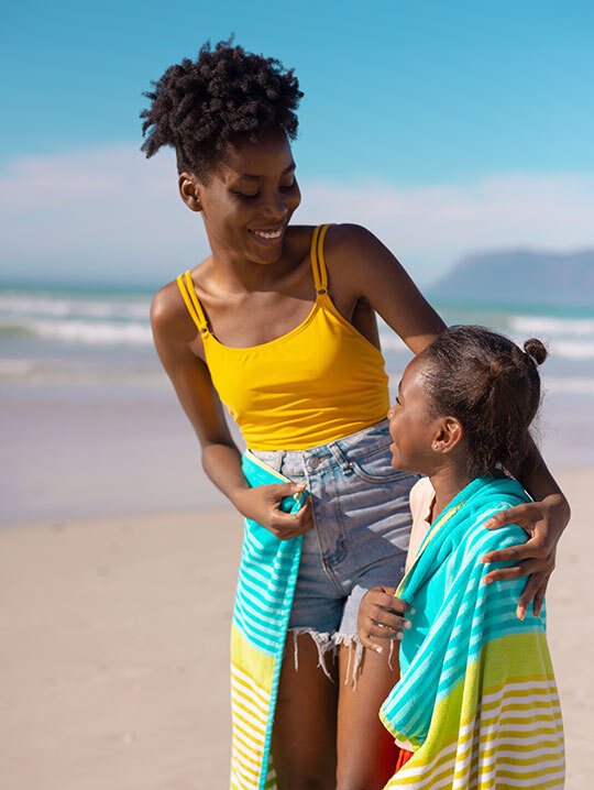 Mutter und Kind stehen in ein Handtuch gewickelt am Strand und lächeln sich an