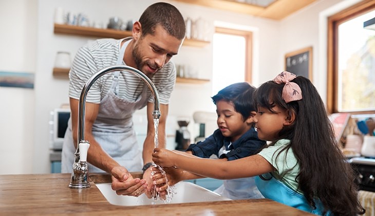 Ein Mann mit einer Kochschürze steht mit zwei Kindern am Waschbecken und alle waschen Hände
