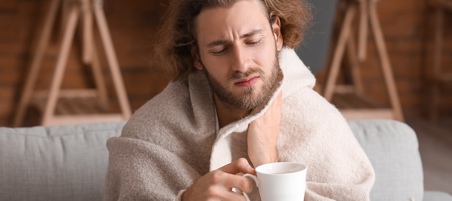 Ein Mann sitzt mit einer Decke auf der Couch, fasst sich den Hals vor Schmerzen und hält eine Teetasse in der Hand