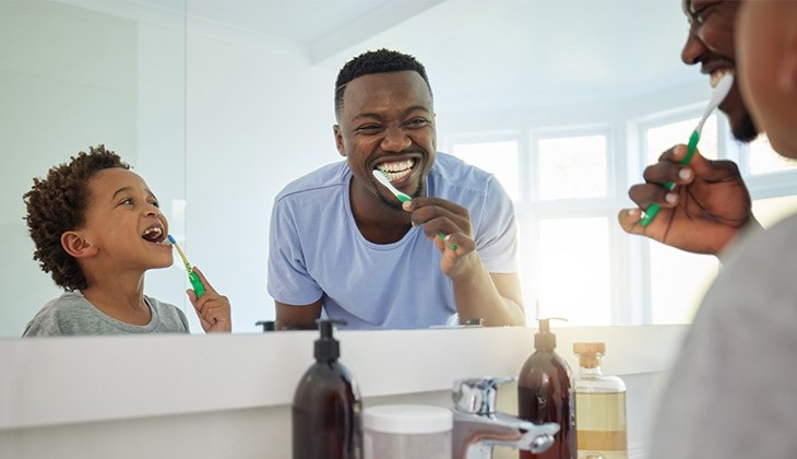 Ein kleines Kind und ein Mann putzen sich vor dem Badezimmerspiegel lächelnd die Zähne.