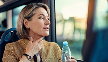 Frau sitzt im Bus, hält eine Wasserflasche und fasst sich an den Hals, als hätte sie Halsschmerzen.