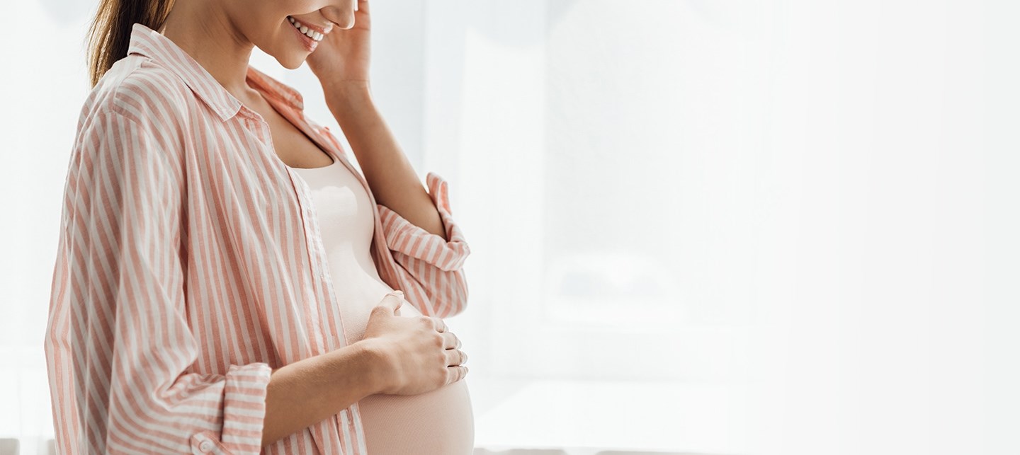 Eine Schwangere Frau hält lächelnd ihre rechte Hand an ihren Bauch