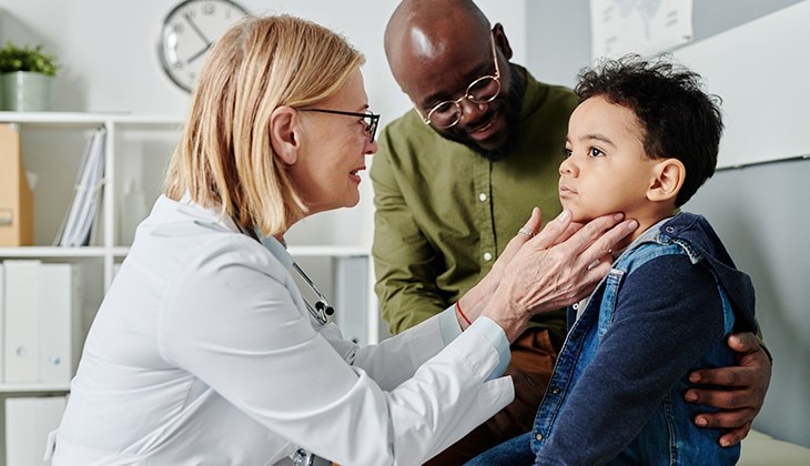 Kind wird von einer Ärztin am Hals untersucht und der Vater sitzt daneben