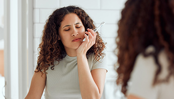 Eine junge Frau hält sich beim Zähneputzen vor dem Badezimmerspiegel mit der Hand die schmerzende Wange. 