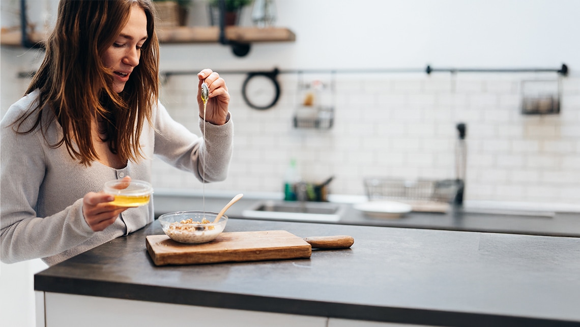 Eine junge Frau steht in einer modernen Küche und gießt mit einem Löffel Honig über eine Schüssel Müsli.