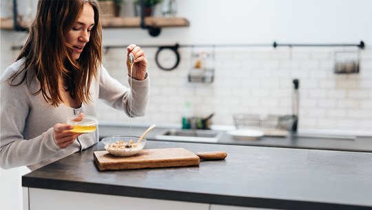 Eine junge Frau steht in einer modernen Küche und gießt mit einem Löffel Honig über eine Schüssel Müsli.