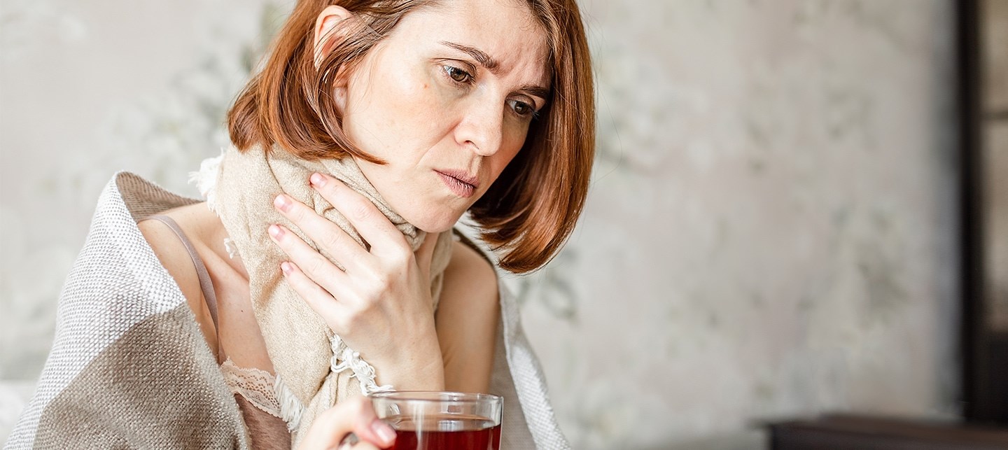 Eine rothaarige Frau mit Schal umgebunden, hält sich den Hals vor Schmerzen und trinkt Tee